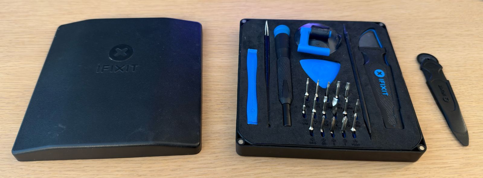 An iFixit Essential Electronics Toolkit open on a wooden desk: the lid on the left, the tray with screwdriver, bits, spudger, tweezers, suction handle and opening pick on the right, and a black iMac opening wheel beside it.