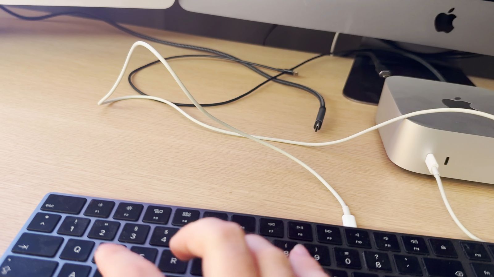 A hand typing on a Magic Keyboard that is connected to the Mac mini with a white USB-C cable; the Mac mini's status light is on.