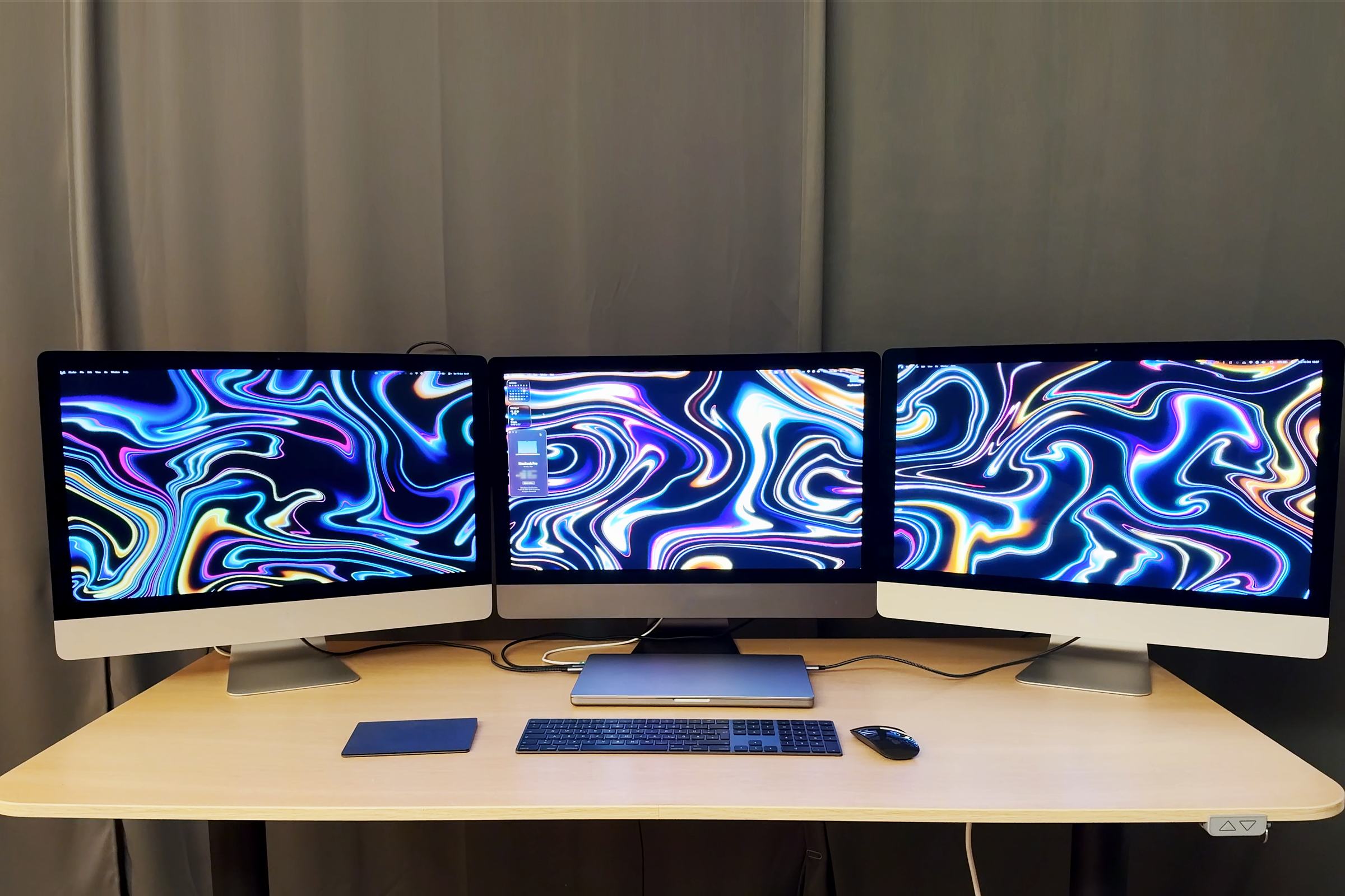 Three 27-inch iMacs side by side with a swirling wallpaper across all three screens, working as displays for the MacBook Pro that sits closed on the desk in front of them, connected by cables.