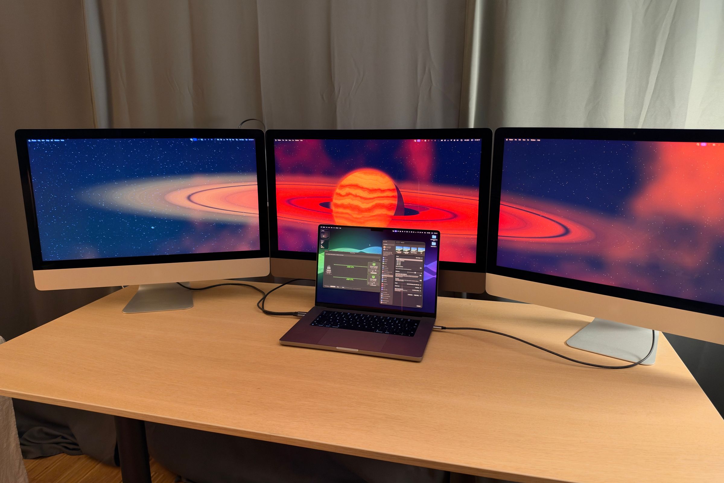 Three 27-inch iMacs side by side on a desk, all showing a ringed-planet wallpaper, with a MacBook Pro in front, connected by cables and showing the DisplayBridge window.