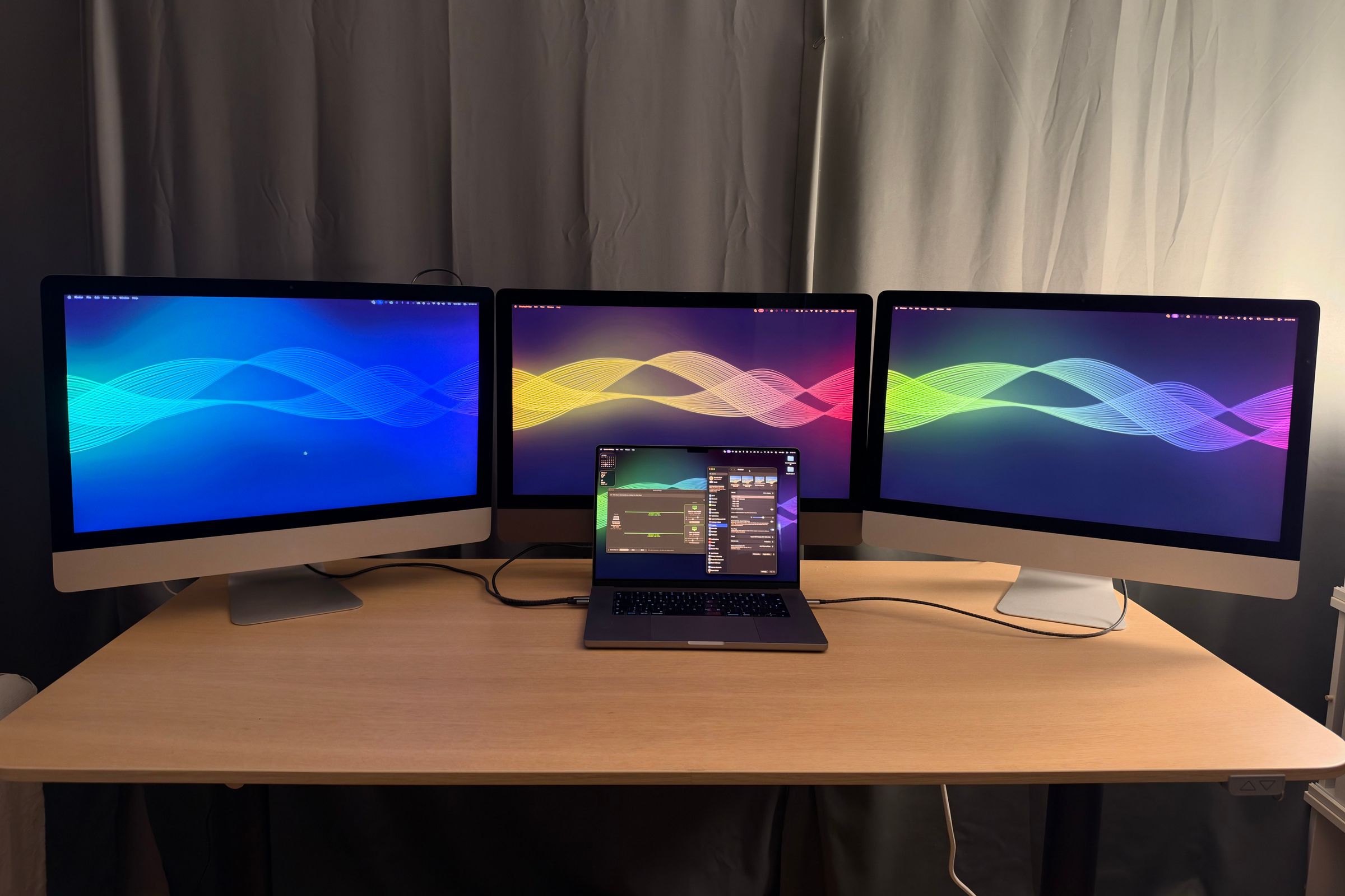 Three 27-inch iMacs with colourful wave wallpapers as extended displays of the MacBook Pro on the desk in front of them.