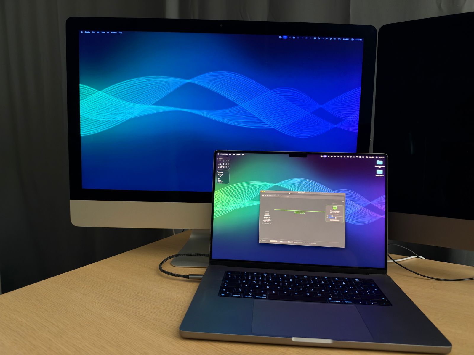 A 27-inch 5K iMac with a blue wave wallpaper as the second display of the MacBook Pro in front of it, which shows the DisplayBridge window; one cable connects them.
