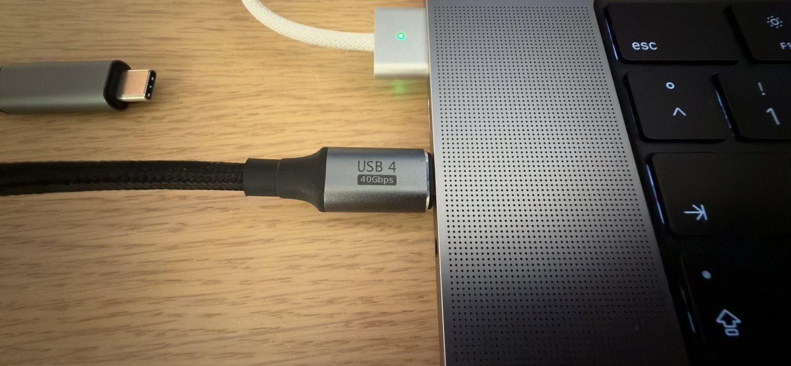Close-up of a black braided cable plugged into the left side of a MacBook Pro; the silver plug is labelled USB 4, 40Gbps. A MagSafe charger is plugged in above it.